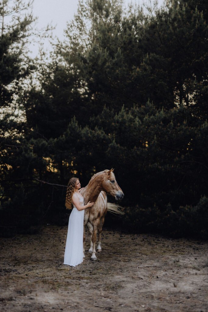 Eine Frau in einem weißen Kleid steht neben einem Pferd im Wald.