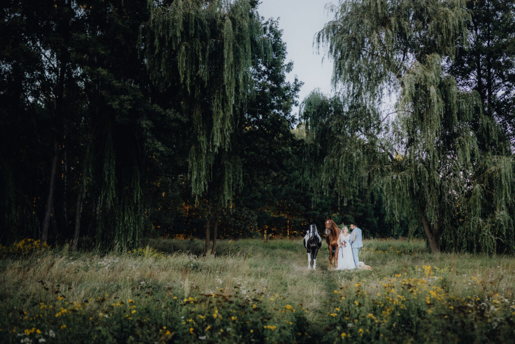 Eine Braut und ein Bräutigam stehen mit ihren Pferden auf einem Feld mit Weidenbäumen.