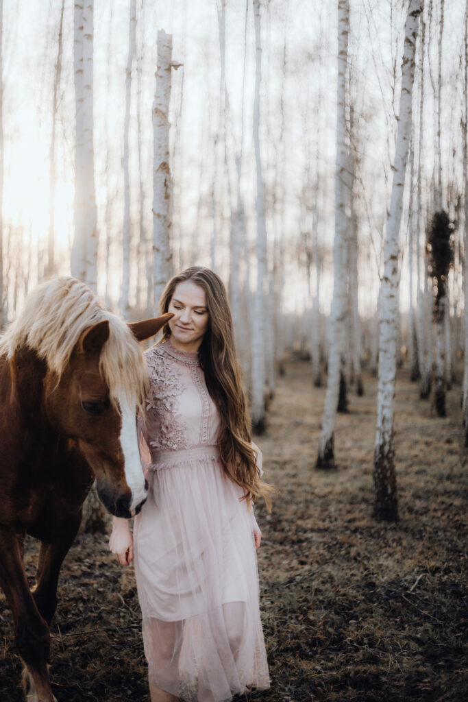 Eine Frau in einem rosa Kleid steht neben einem Pferd im Wald.