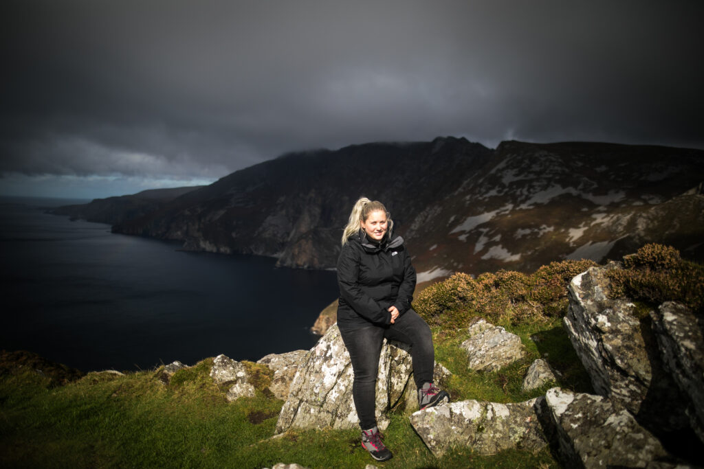 Gina Wetzler genießt einen seltenen Sonnenstrahl auf einer sonst düsteren Küstenlandschaft in Irland.
